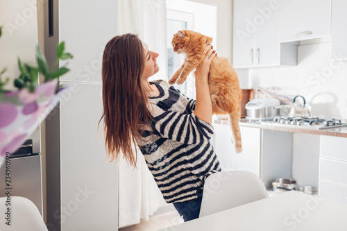 Young woman playing with cat in kitchen at home. Girl holding and raising red cat