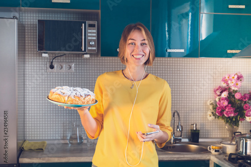 Smiling woman listen music at the headphones and demonstration hot homemade pie. Blue kitchen on the background