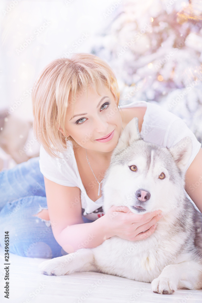 Beautiful blonde woman sits with husky dog near christmas tree.