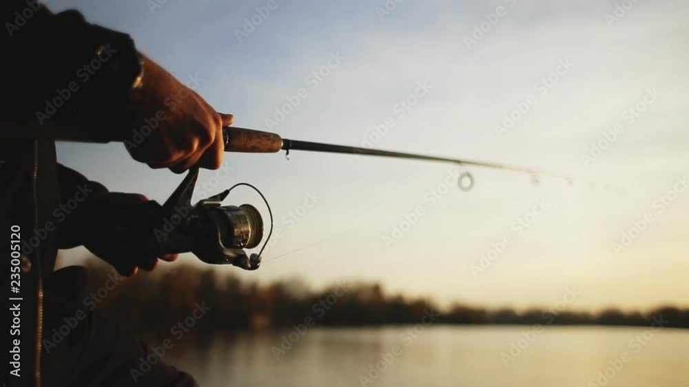 Fishing on the lake at sunset. A young man is fishing for spinning against the background of the setting sun. Silhouette of a fisherman. Hands close up.