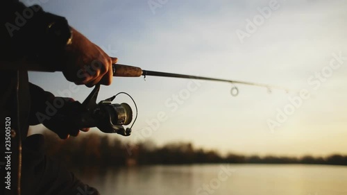 Fishing on the lake at sunset. A young man is fishing for spinning against the background of the setting sun. Silhouette of a fisherman. Hands close up.