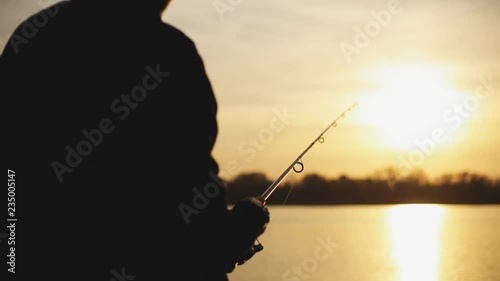 Fishing on the lake at sunset. A young man is fishing for spinning against the background of the setting sun. Silhouette of a fisherman. Hands close up.
