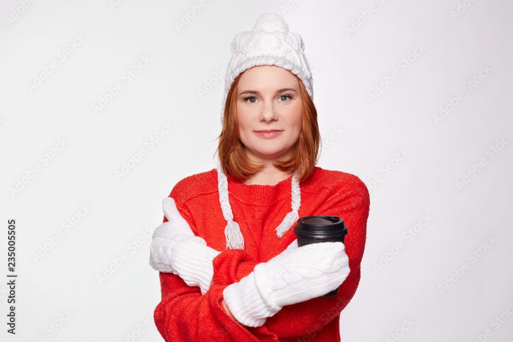 Christmas winter time concept. Studio shot of creative good-looking European freckled woman with red hair wearing trendy hat and stylish red hoodie, smiling gently, holding cup with hot beverage. Christmas winter time concept. Studio shot of creative good-looking European freckled woman with red hair wearing trendy hat and stylish red hoodie, smiling gently, holding cup with hot beverage.