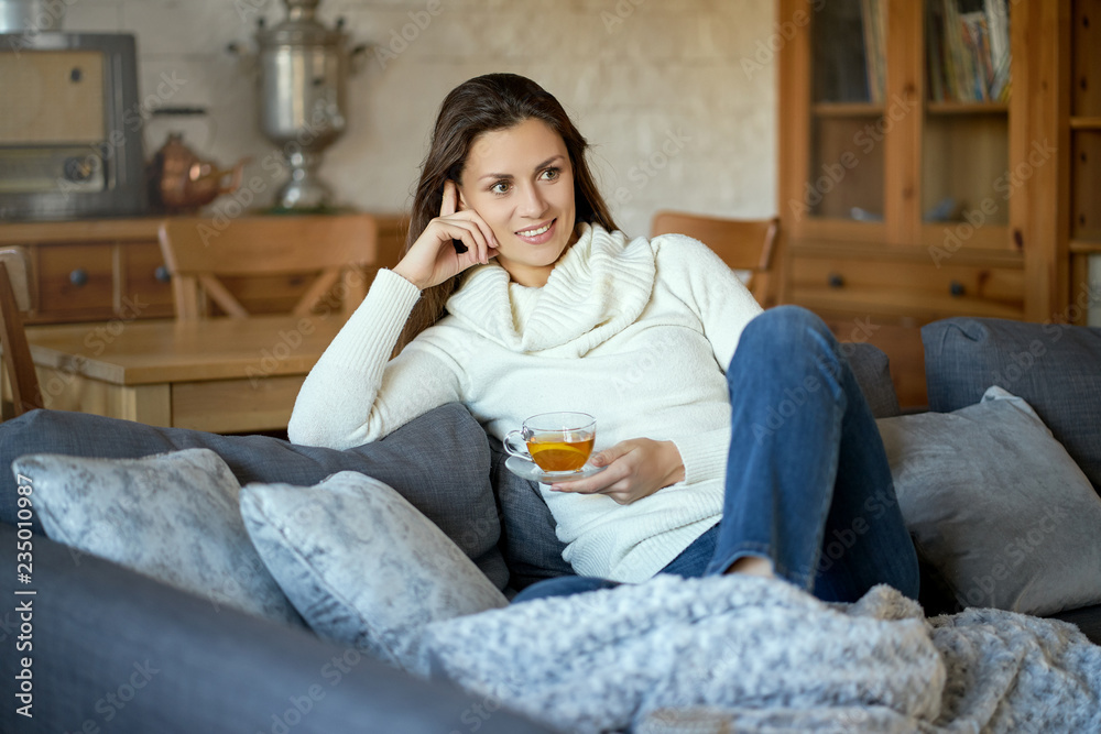 attractive young woman in sweater relaxes on a gray sofa at home and drinks tea with lemon attractive young woman in sweater relaxes on a gray sofa at home and drinks tea with lemon