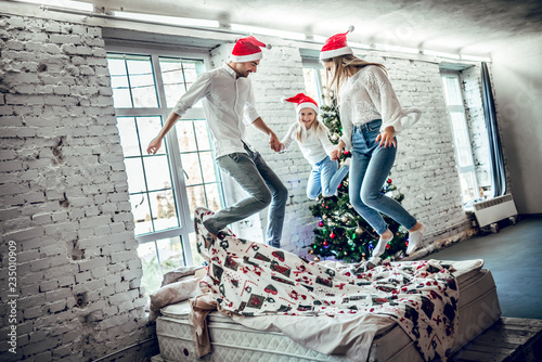 Funny moments at this holiday. Houng family holding hands and cheerfully jumping on the bed. Christmas tree in background. photo