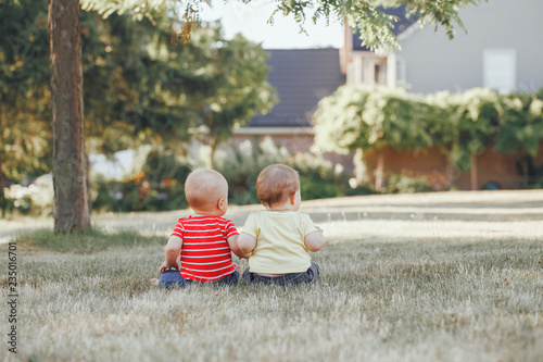 Two adorable little Caucasian babies sitting together in field meadow outside.View from back behind. Little happy children in summer park on sunset. Talking communication with friend concept photo