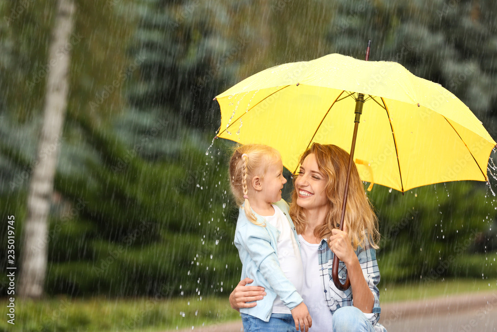 Fototapeta premium Portrait of happy mother and daughter with yellow umbrella in park on rainy day. Space for text