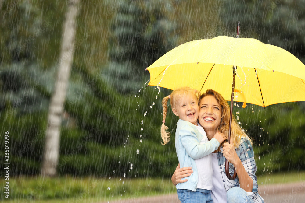Obraz premium Portrait of happy mother and daughter with yellow umbrella in park on rainy day. Space for text
