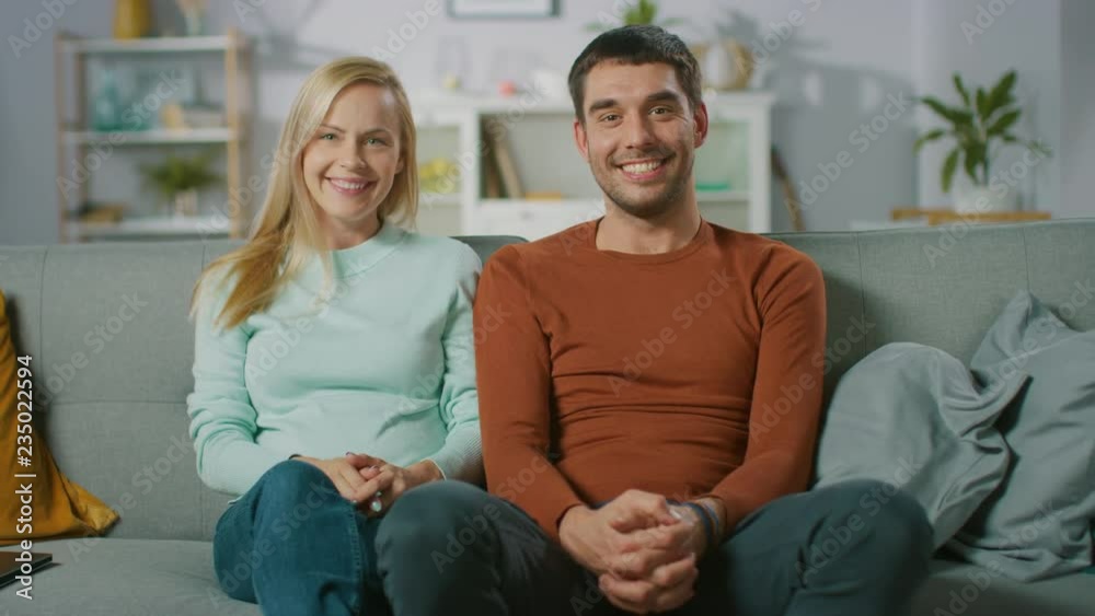 Cute Young Couple Sits on a Sofa at Home, They Look Lovingly at Each Other and Then Smile at Camera. Portrait of Beautiful Happy Young Couple. Cute Young Couple Sits on a Sofa at Home, They Look Lovingly at Each Other and Then Smile at Camera. Portrait of Beautiful Happy Young Couple.