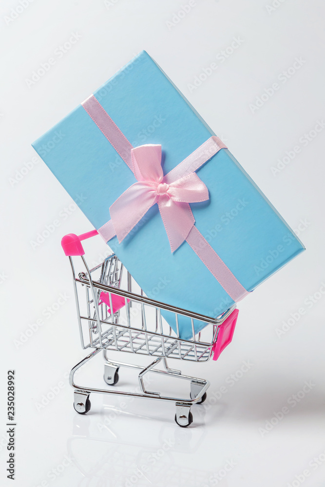 Small supermarket grocery push cart for shopping toy with blue gift box isolated on white background. Sale buy mall market shop consumer concept. Copy space Small supermarket grocery push cart for shopping toy with blue gift box isolated on white background. Sale buy mall market shop consumer concept. Copy space