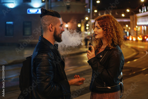 Couple in love on date. Boyfriend talking to girlfriend. She smokes an electronic cigarette and looks at him. They walk on streets of night city. Wet asphalt after rain.