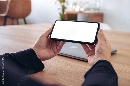 Mockup image of hands holding black mobile phone with blank white screen horizontally for watching with laptop on table in office