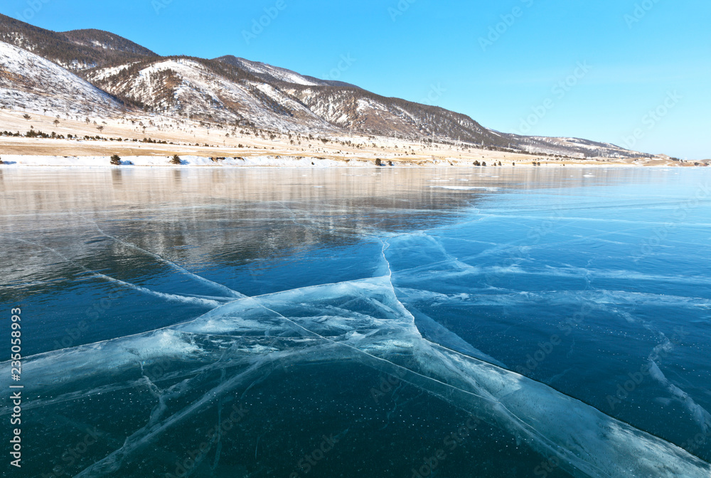 Naklejka premium Lake Baikal in the winter. View of the snow-covered coast of the Small Sea Strait from smooth blue ice with cracks on a sunny February day