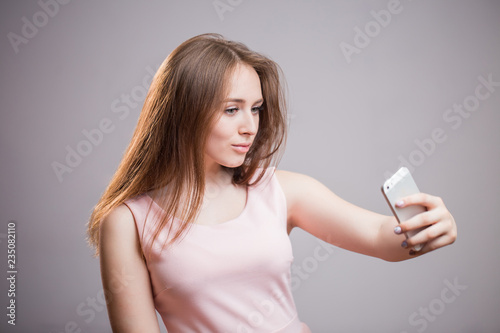 Beautiful young woman in pink dress taking selfie on her smartphone and smiles on grey background
