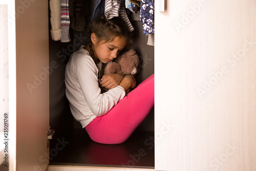 Domestic violence and abused concept. Unhappy and scared child hiding in the wardrobe and sitting there down in the mouth with Teddy in her hands