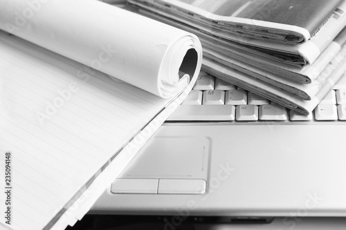 Business concept. Stack of fresh morning newspapers with business news and open paper notebook with blank sheets on keyboard of laptop, side view 