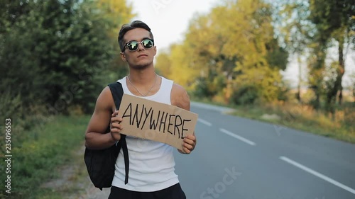 A tourist hitchhiker walks along the road with a backpack on his shoulder and sign Anywhere. Traveler is trying to stop the car