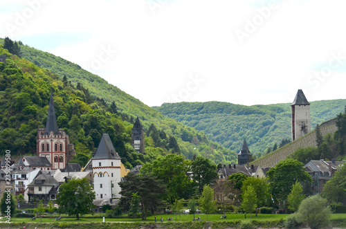A small village of traditional houses is by a river in Germany. Castle, church and granary towers rise above the buildings. The surrounding hills are covered with forest, with a vineyard is on one sl