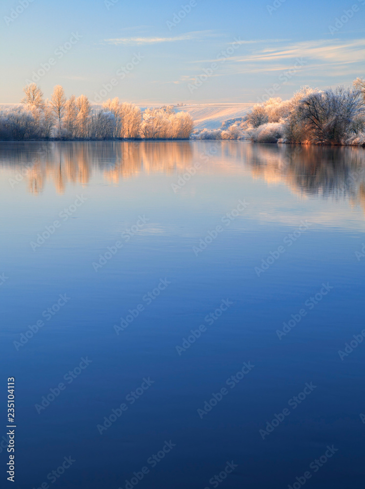 Winter Landscape Reflecting in River at Sunrise, Trees covered by hoarfrost and snow, blue sky