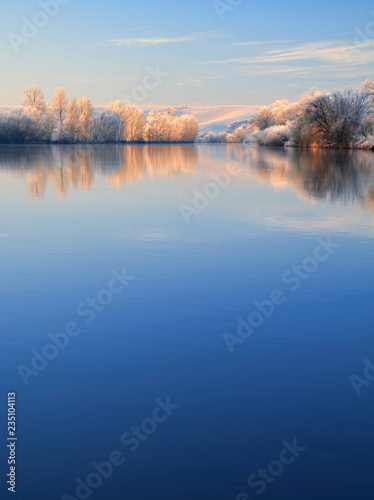 Winter Landscape Reflecting in River at Sunrise, Trees covered by hoarfrost and snow, blue sky