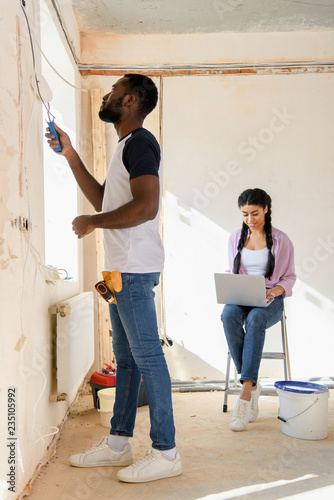 side view of african american man painting wall while his girlfriend using laptop behind during renovation of home