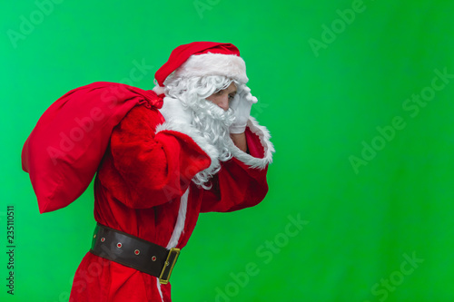 Christmas. Kind and tired Santa Claus in white gloves carries a red bag with gifts over his shoulder. Isolated on green background