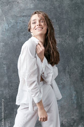 Smiling brunette female model posing in white shirt, top and pants. Studio shot, grey background © monchak