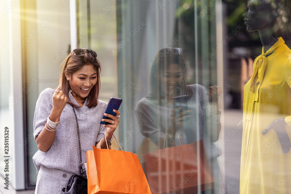 Asian woman cheerful when using the smart mobile phone for check online shopping order is completed with clothes beside the glassess in store shop with happy action at department center