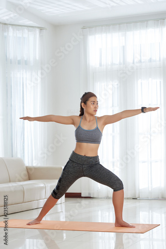 Young muscular Asian woman in activewear standing in Warrior yoga pose during morning practice at home