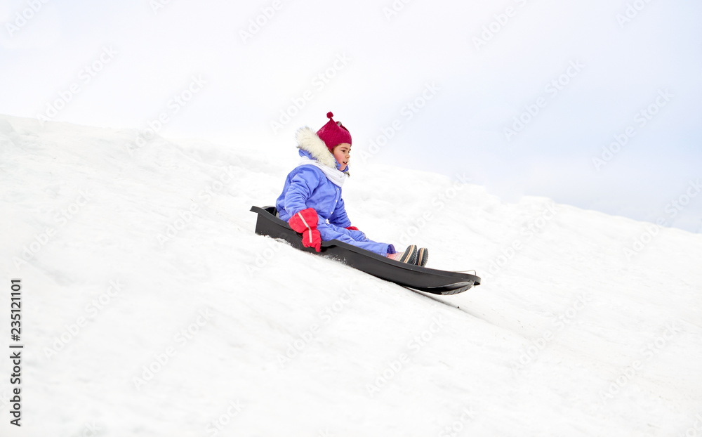 childhood, sledging and season concept - happy little girl sliding on sled down snow hill outdoors in winter