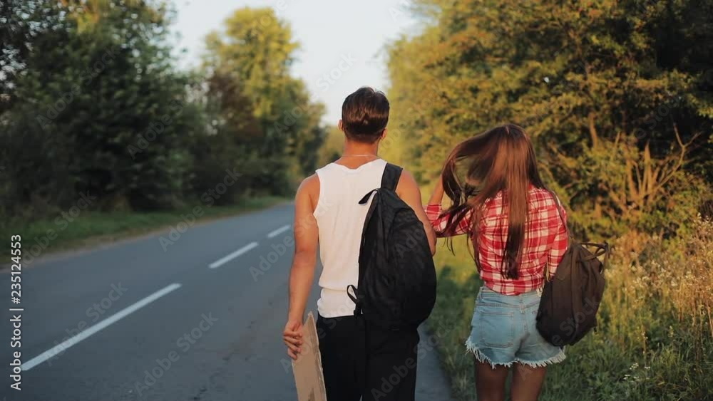 Couple On Vacation Hitchhiking Along Road and Holding Adventure Sign. Summer time. Hitchhiking, tourists, adventures concept Couple On Vacation Hitchhiking Along Road and Holding Adventure Sign. Summer time. Hitchhiking, tourists, adventures concept
