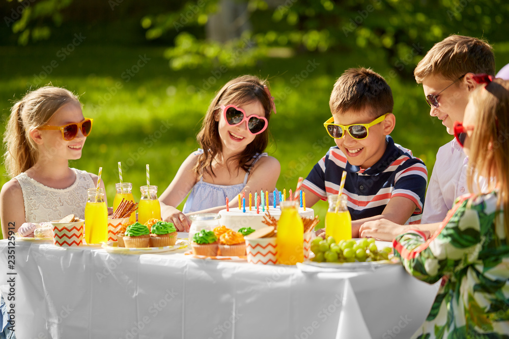holidays, childhood and celebration concept - happy kids with candles on birthday cake sitting at table at summer garden party holidays, childhood and celebration concept - happy kids with candles on birthday cake sitting at table at summer garden party