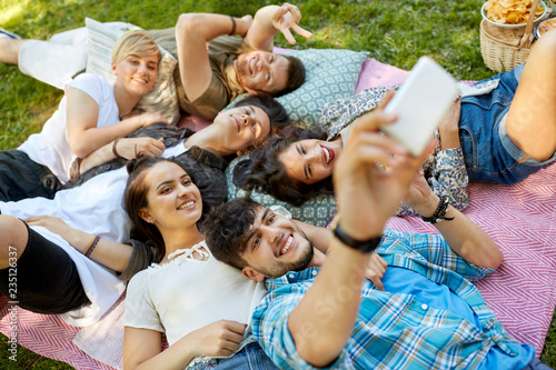 friendship  leisure  technology and people concept - group of friends chilling on picnic blanket at summer park and taking selfie by smartphones
