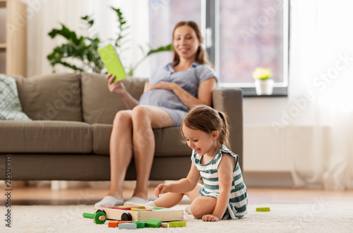 childhood and people concept - happy three years old baby girl playing with toy blocks and pregnant mother reading book at home
