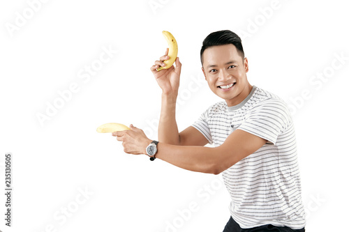 Portrait of young handsome Asian man holding bananas like pistols and smiling at camera cheerfully on white background