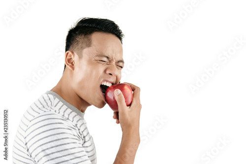 Waist-up portrait of young Asian man wincing painfully while eating fresh red apple against white background, copy space