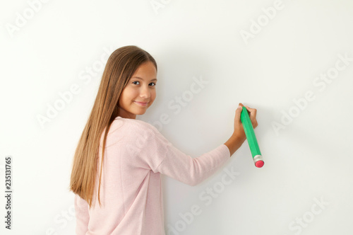 A young schoolgirl is writing with a pen in his hand in front of a white wall.