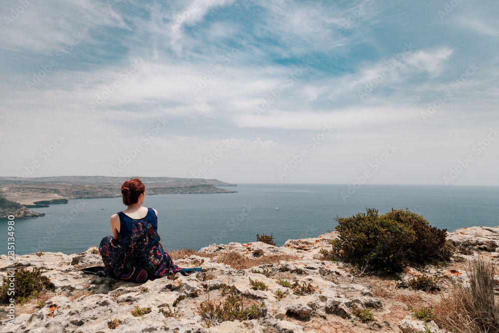 Young redhead woman with a topknot sitting on the edge of a cliff looking far away the sea Young redhead woman with a topknot sitting on the edge of a cliff looking far away the sea