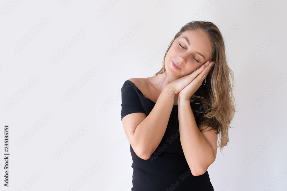 Content pretty Caucasian girl showing sleep symbol and smiling. Young woman in black dress closing eyes and putting head on hands. Sweet dreams concept