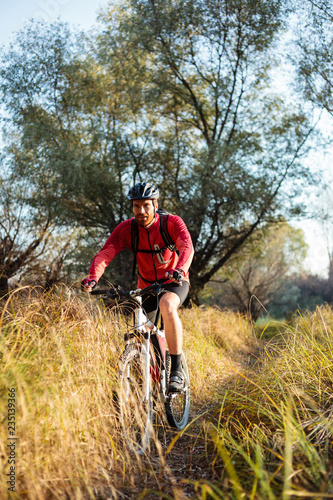 Smiling young bearded man in red long sleeve cycling jersey riding mountain bike along a path through tall grass. Low angle front view. © Ivan