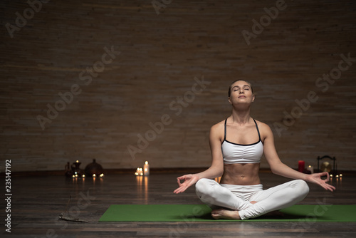 Calm relaxed young lady sitting on the yoga mat with her legs crossed and meditating with candles on the background
