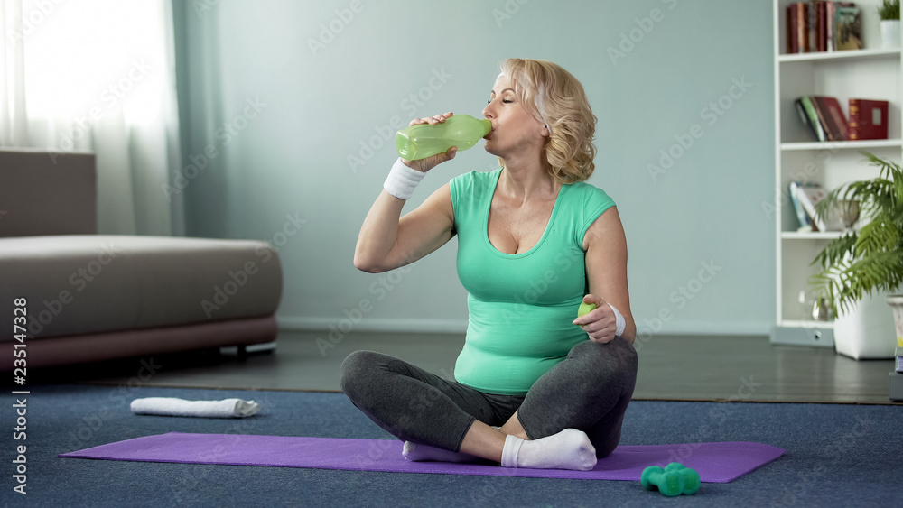 Athletic senior woman sitting on floor and drinking water after workout, health Athletic senior woman sitting on floor and drinking water after workout, health