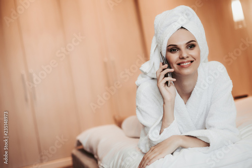 Concept of relaxation and communication after spa. Waist up portrait of young smiling lady in bathrobe talking by phone while lying on bed. Copy space on left