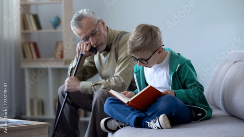 Boy in glasses reading book for grandfather, old man falling asleep on cosy sofa
