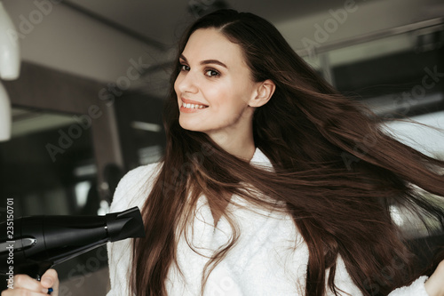 Concept of haircare and treatment after shower. Close up portrait of smiling beautiful woman enjoying while drying hair with blowdryer at home bathroom