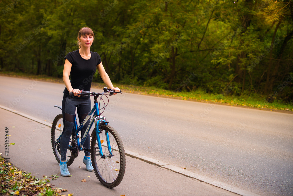 Fototapeta premium Young beautiful athletic girl in sportswear sitting on a blue bike on the sidewalk. Trees on the background.