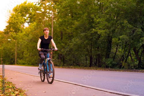 young beautiful girl in sportswear rides a blue bike on the sidewalk. Trees on the background. © Павел Страхов