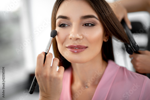Cheerful emotional young woman visiting professional beauty salon and smiling while her beautician putting rouge on her face