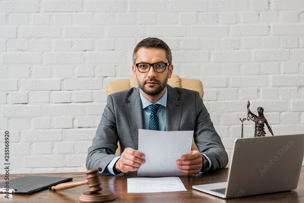 serious lawyer in eyeglasses working with papers and looking at camera in office serious lawyer in eyeglasses working with papers and looking at camera in office