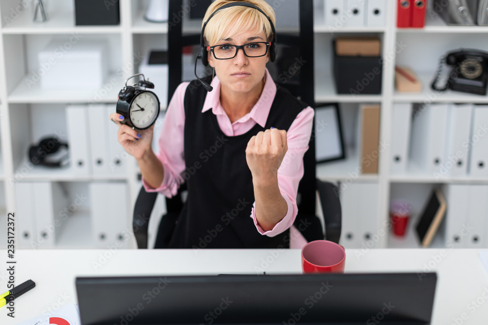 A young girl sitting at a computer table and holding an alarm clock.